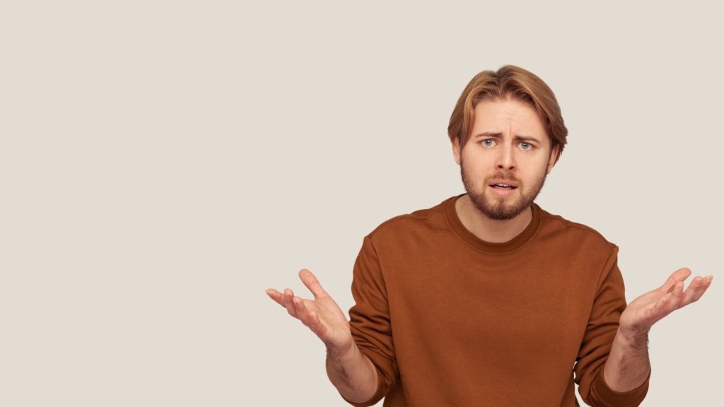 Confused man shrugging with open hands, representing accounting or financial questions, in a brown sweatshirt against a plain beige background.